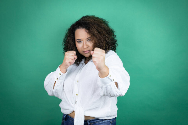 Chica afroamericana joven con camisa blanca sobre fondo verde puñetazo en el puño para luchar, ataque agresivo y enojado, amenaza y violencia - Foto, imagen