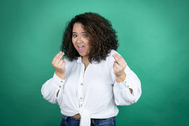 Jeune fille afro-américaine portant une chemise blanche sur fond vert faisant geste d'argent avec les mains, demandant le paiement du salaire, entreprise millionnaire - Photo, image