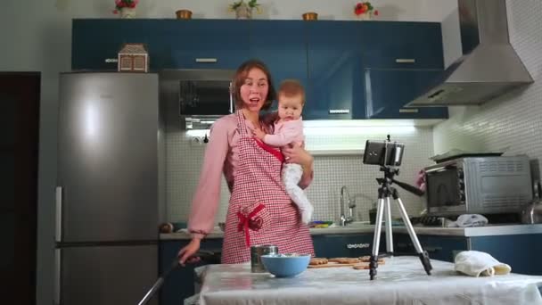 Una madre sostiene a un niño en sus brazos, aspira la cocina y graba una transmisión en vivo en su teléfono inteligente. El concepto de multitarea - Imágenes, Vídeo