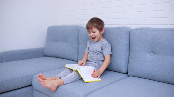 Mały chłopiec w szarych ciuchach, siedzący na szarej sofie i uczący się czytać żółtą książkę. Uwielbiam czytać, spędzać czas z rodziną. Samoizolacja, uczenie się na odległość. - Zdjęcie, obraz