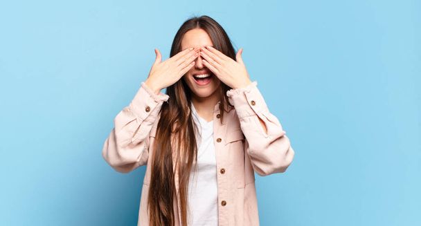 young pretty casual woman smiling and feeling happy, covering eyes with both hands and waiting for unbelievable surprise - Фото, изображение