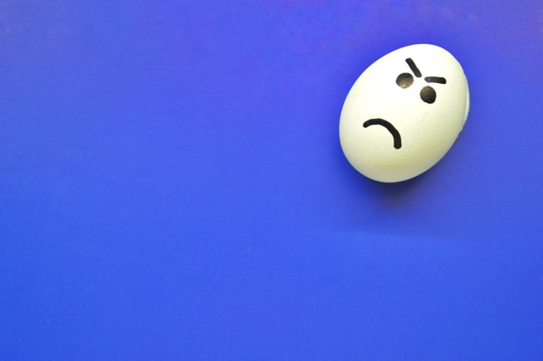 Uovo bianco con volto arrabbiato dipinto su sfondo blu. - Foto, immagini