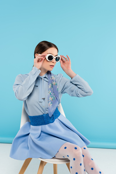 elegant woman adjusting sunglasses while sitting on blue background - Fotografie, Obrázek