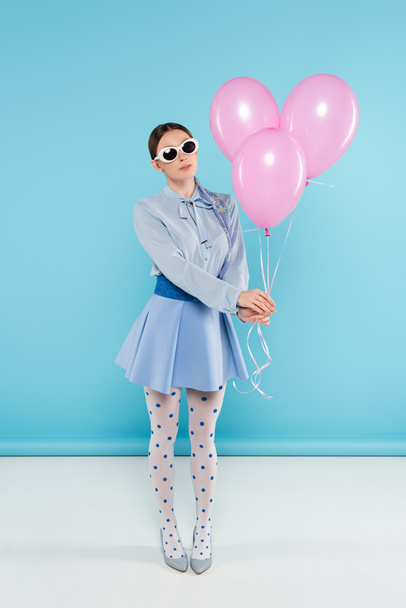 extravagantní žena v puntíkové punčocháče a sluneční brýle drží růžové balónky na modrém pozadí - Fotografie, Obrázek