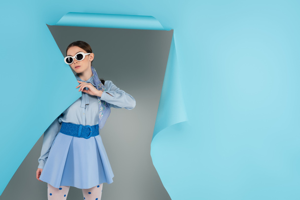 young fashionable woman in sunglasses near hole in blue paper on grey background - Photo, Image