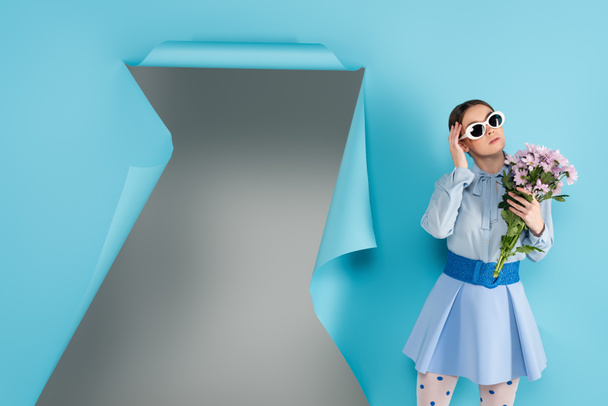 fashionable woman in eyeglasses holding flowers near hole in blue paper with grey background - Photo, Image