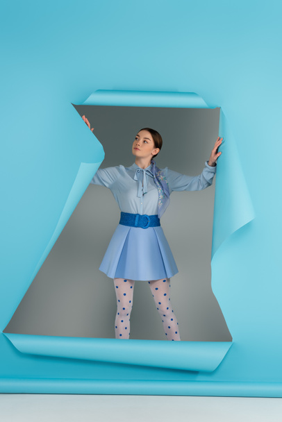mujer glamour en medias punteadas de pie cerca del agujero en papel azul sobre fondo gris - Foto, imagen