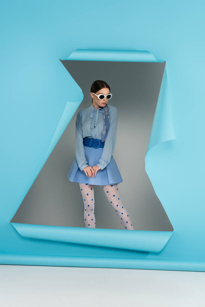 glamour woman in polka dot tights and sunglasses posing near blue ripped paper on grey background - Photo, Image