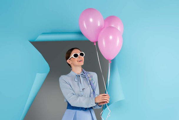 Gözlüklü mutlu kadın gri arka planda mavi yırtık kağıdın yanında pembe balonlar tutuyor. - Fotoğraf, Görsel
