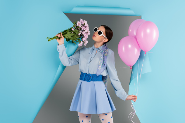 stylová žena pózující s růžovými květy a balónky v blízkosti modré roztrhl papír na šedém pozadí - Fotografie, Obrázek