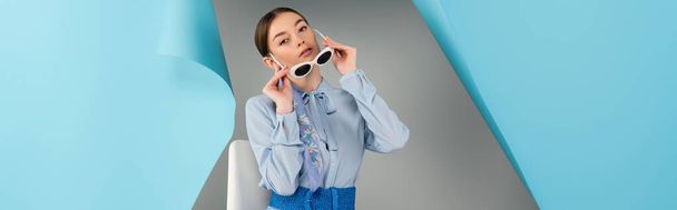 stylish woman taking off sunglasses near hole in blue paper on grey background, banner - Photo, Image