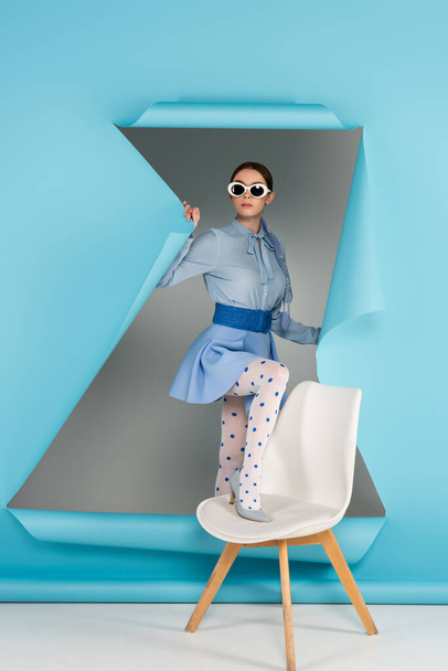 Lekeli tayt ve güneş gözlüğü takmış zarif bir kadın gri arka planda mavi yırtık kağıdın yanındaki sandalyeye basıyor. - Fotoğraf, Görsel