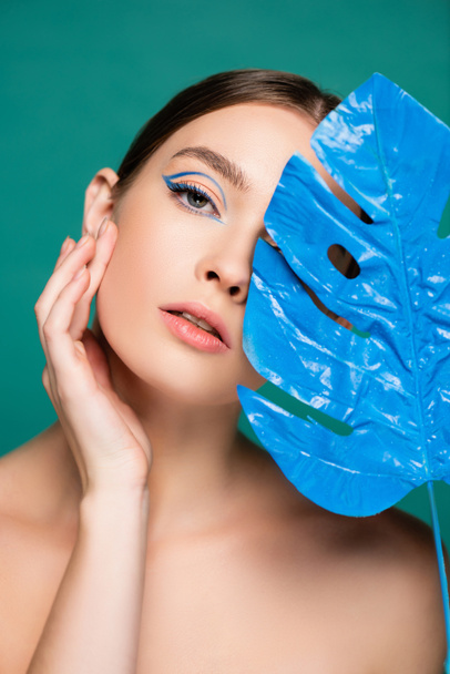 charming woman touching face while looking at camera near blue wet leaf isolated on green - Photo, Image