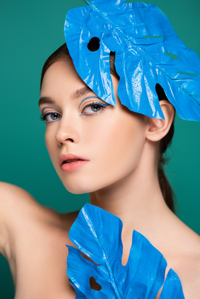 femme sensuelle avec maquillage créatif regardant la caméra près de feuilles brillantes bleues isolées sur vert - Photo, image