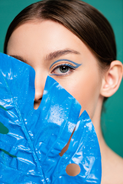 retrato de jovem mulher obscurecendo rosto com folha molhada azul isolado em verde - Foto, Imagem