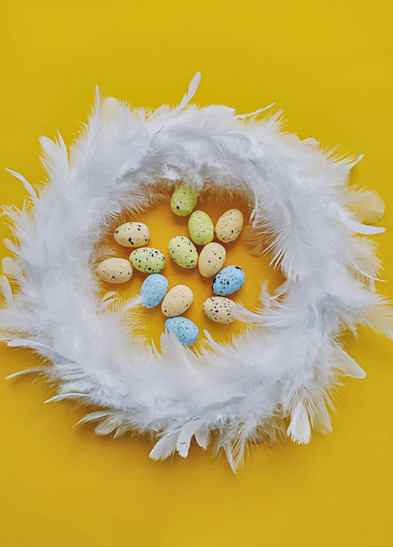White feather wreath on yellow background with mini artificial eggs, top view. Easter decoration concept - Photo, Image