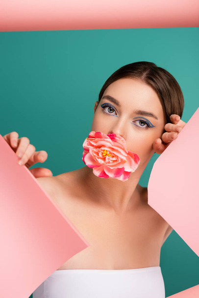 mooie vrouw met pioenroos bloem in de mond kijken naar camera in de buurt gat in roze papier geïsoleerd op groen - Foto, afbeelding