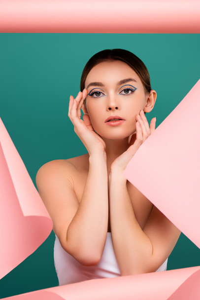 young woman with creative makeup touching face near hole in pink paper isolated on green - Foto, Bild
