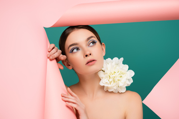 young woman with white peony on shoulder looking out hole in pink paper isolated on green - Фото, зображення