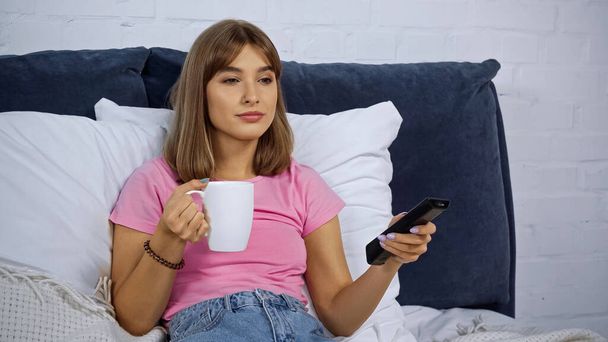 молодая женщина держит чашку и пульт дистанционного управления во время просмотра фильма в спальне - Фото, изображение