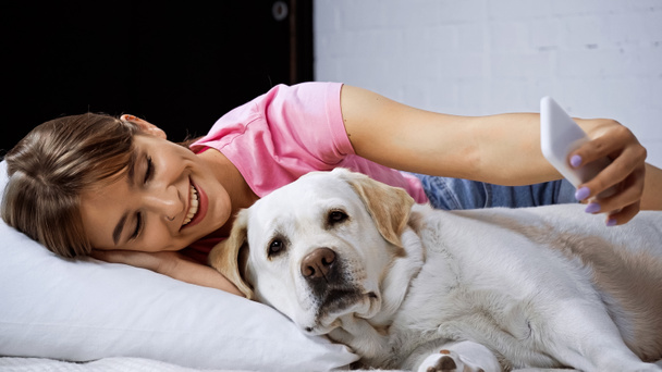 happy young woman lying on bed and taking selfie with golden retriever - Photo, Image
