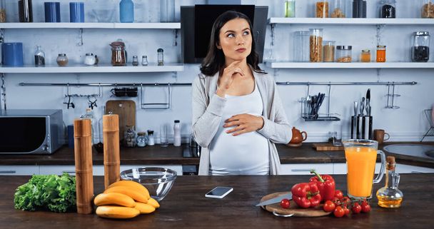 Pensive έγκυος γυναίκα στέκεται κοντά smartphone και φρέσκα συστατικά στην κουζίνα  - Φωτογραφία, εικόνα