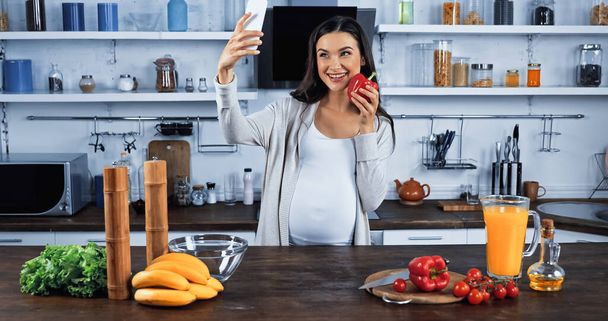 Mulher grávida tomando selfie enquanto segurando pimentão perto de comida na cozinha  - Foto, Imagem