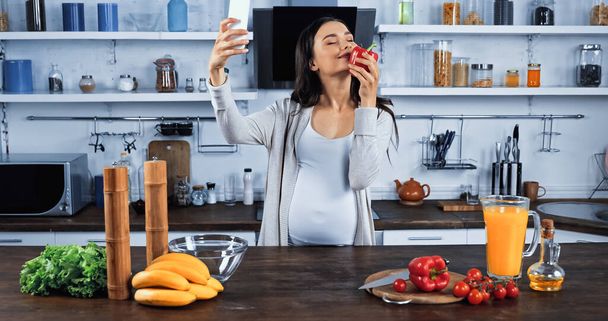 Mujer embarazada oliendo pimiento y tomando selfie en el teléfono inteligente en la cocina  - Foto, Imagen