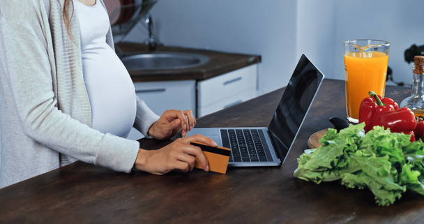 Widok ciężarnej kobiety trzymającej kartę kredytową w pobliżu laptopa i świeżego jedzenia w kuchni  - Zdjęcie, obraz