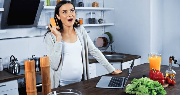 Excited pregnant woman holding credit card near laptop and food in kitchen  - 写真・画像