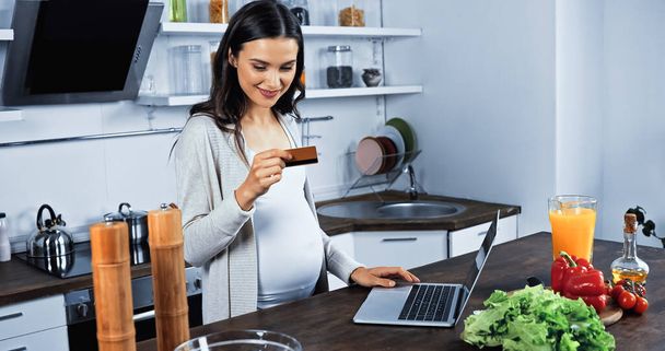 Sourire femme enceinte regardant la carte de crédit près de l'ordinateur portable et des ingrédients sur la table de cuisine  - Photo, image