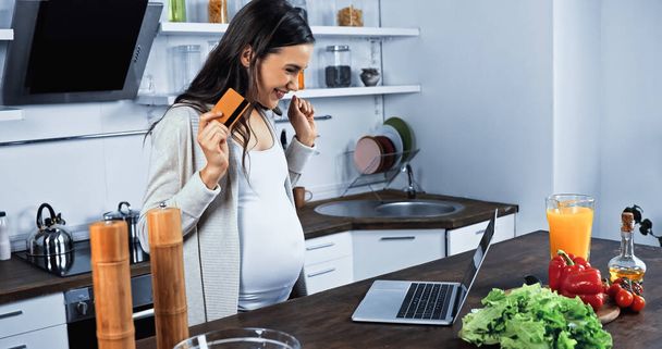 Veselá těhotná žena s kreditní kartou při pohledu na notebook v blízkosti potravin na kuchyňském stole  - Fotografie, Obrázek