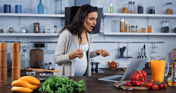 Excited pregnant woman having video call on laptop near fresh food in kitchen  - Photo, Image