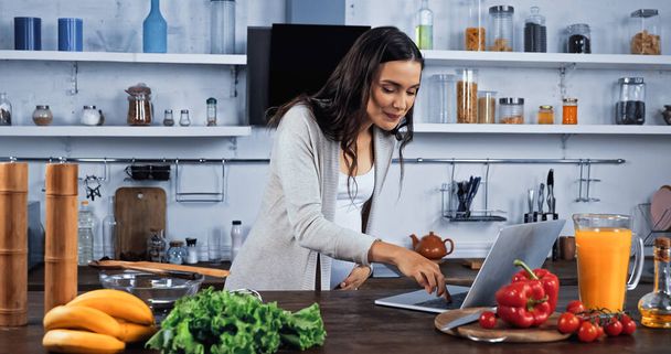 Pregnant woman using laptop near fresh ingredients in kitchen  - 写真・画像