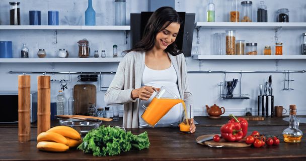 Smiling pregnant woman pouring orange juice near organic vegetables in kitchen  - Photo, Image