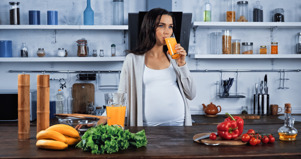 Pregnant woman drinking orange juice near organic food in kitchen  - Photo, Image
