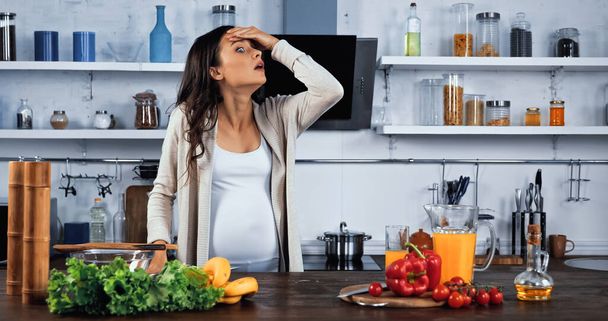 Hämmästynyt raskaana oleva nainen seisoo lähellä tuoretta ruokaa ja appelsiinimehua keittiön pöydällä  - Valokuva, kuva