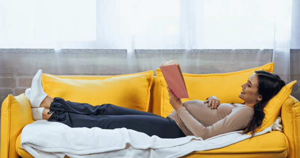 Szczęśliwa kobieta w ciąży odpoczywa z książką na sofie w domu  - Zdjęcie, obraz