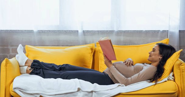 Sonriendo mujer embarazada leyendo libro mientras se relaja en el sofá  - Foto, imagen