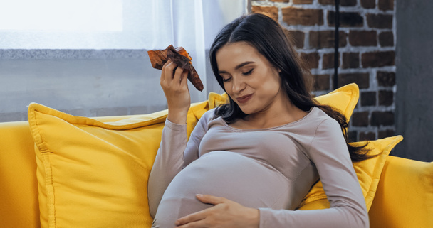 Feliz embarazada comiendo magdalena en el sofá  - Foto, imagen