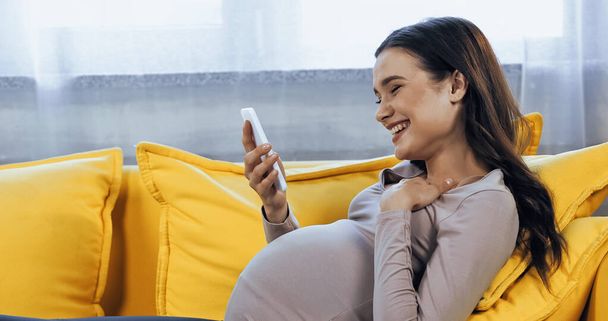 Χαμογελώντας έγκυος γυναίκα κρατώντας smartphone σε φωτεινό κίτρινο καναπέ  - Φωτογραφία, εικόνα