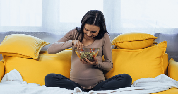 Schwangere hält Schüssel mit frischem Salat auf gelbem Sofa  - Foto, Bild