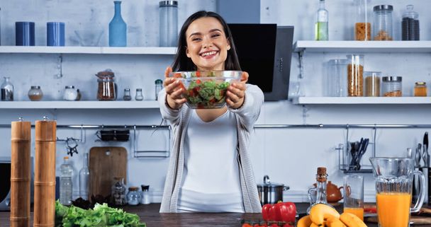Улыбающаяся женщина показывает миску салата рядом со свежей едой на кухонном столе  - Фото, изображение