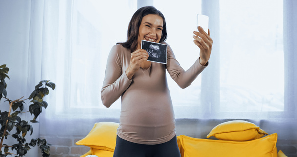 Cheerful pregnant woman showing ultrasound scan of baby during video call  - Фото, изображение