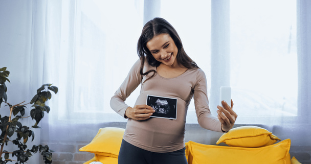 Smiling pregnant woman holding ultrasound scan of baby during video chat  - Fotoğraf, Görsel