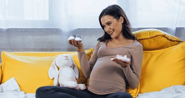 Mujer embarazada sonriente mirando botines de bebé cerca de juguete suave en el sofá  - Foto, Imagen