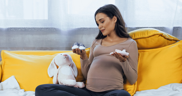 Femme enceinte tenant des chaussons de bébé près de jouet doux à la maison  - Photo, image