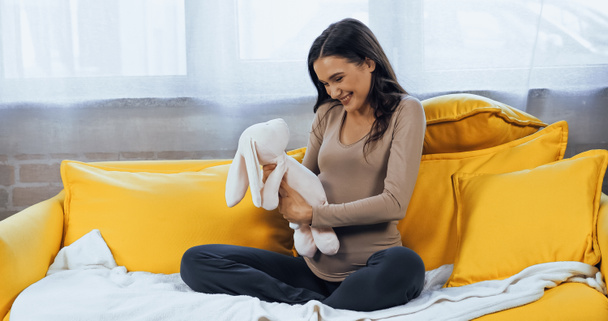 Vidám terhes nő kezében puha játék a nappaliban  - Fotó, kép
