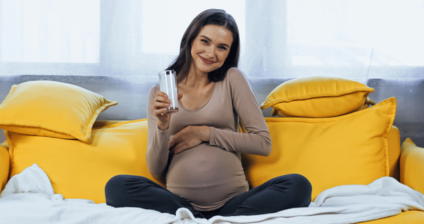 Boldog terhes nő kezében pohár tej sárga kanapé otthon  - Fotó, kép