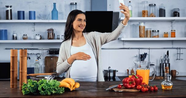 Улыбающаяся беременная женщина делает селфи возле свежей еды на кухне  - Фото, изображение